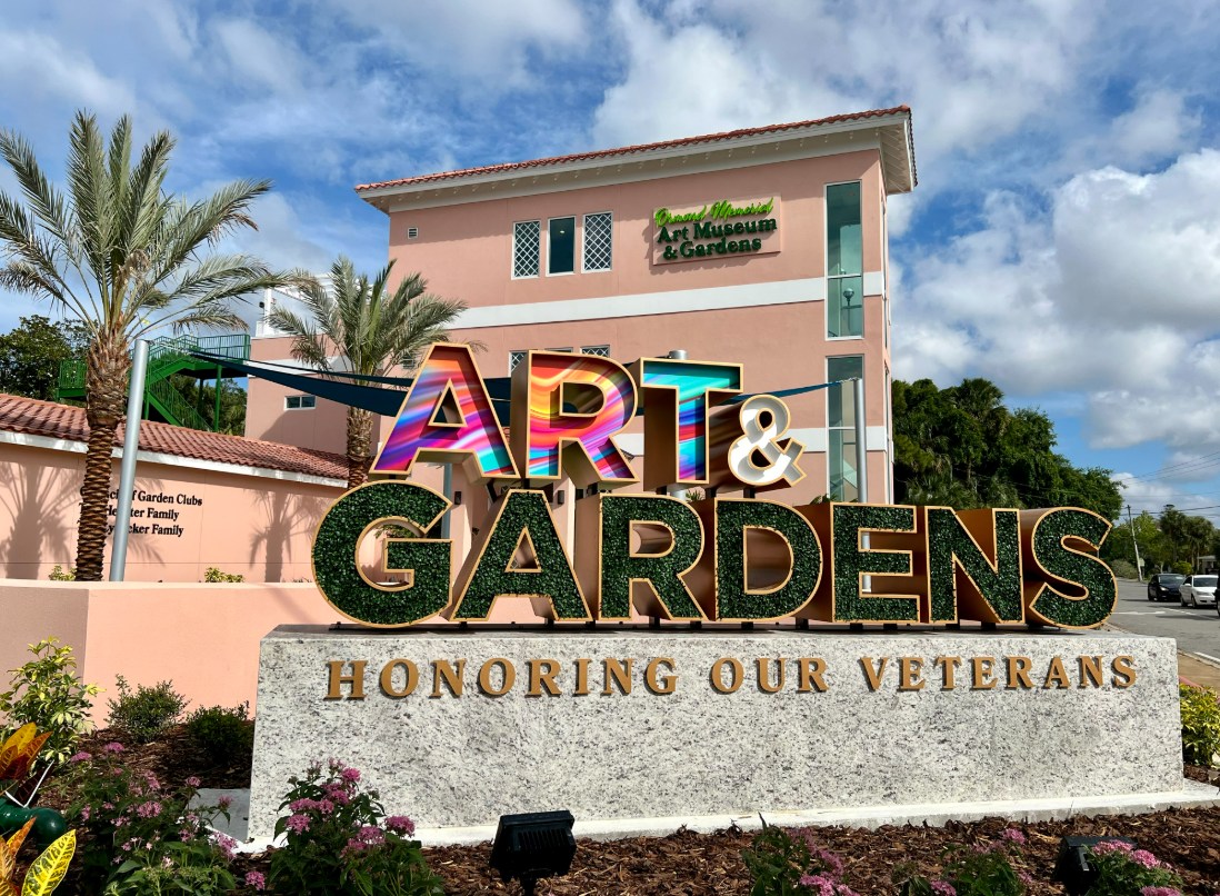 The Art & Gardens sign reading "Honoring Our Veterans" in front of the pink Ormond Memorial Art Museum and Gardens building, framed by palm trees.