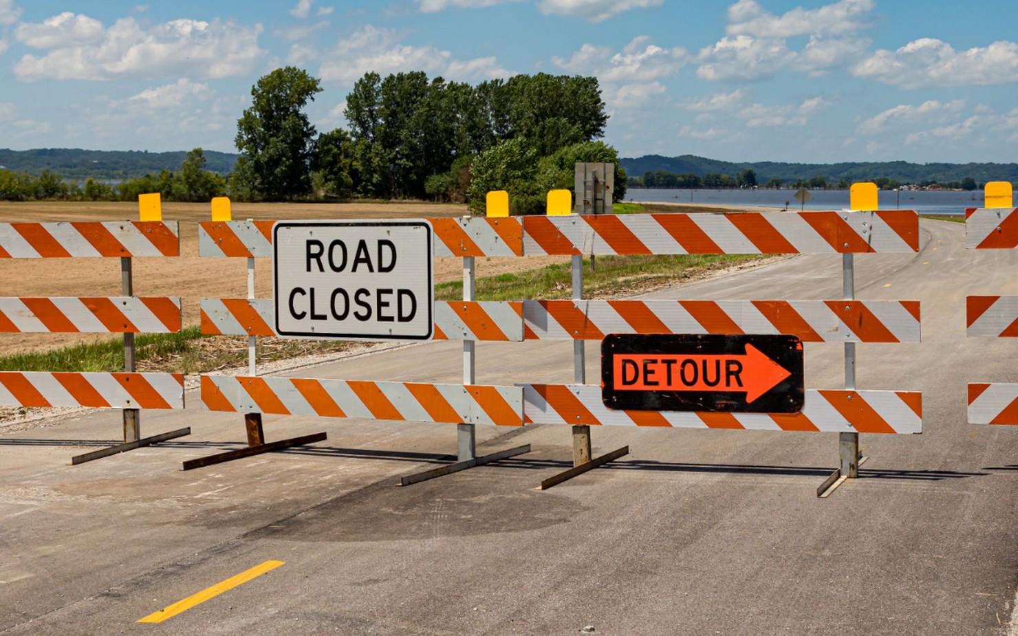Orange and white striped road barricades with a Road Closed sign and an orange Detour arrow blocking a road.