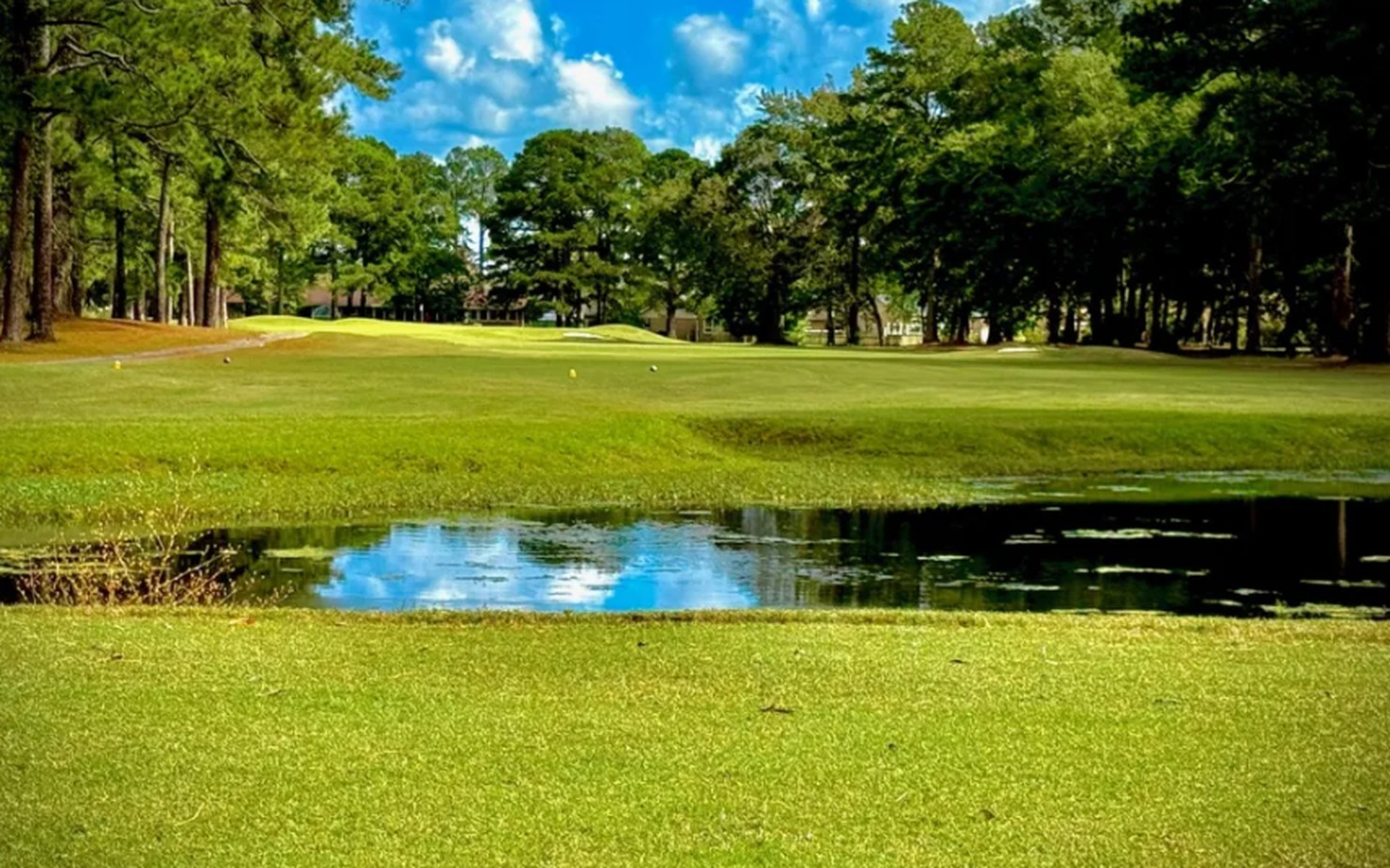 A green fairway at River Bend Golf Course lined with tall pines, with a pond reflecting the blue sky in the foreground.