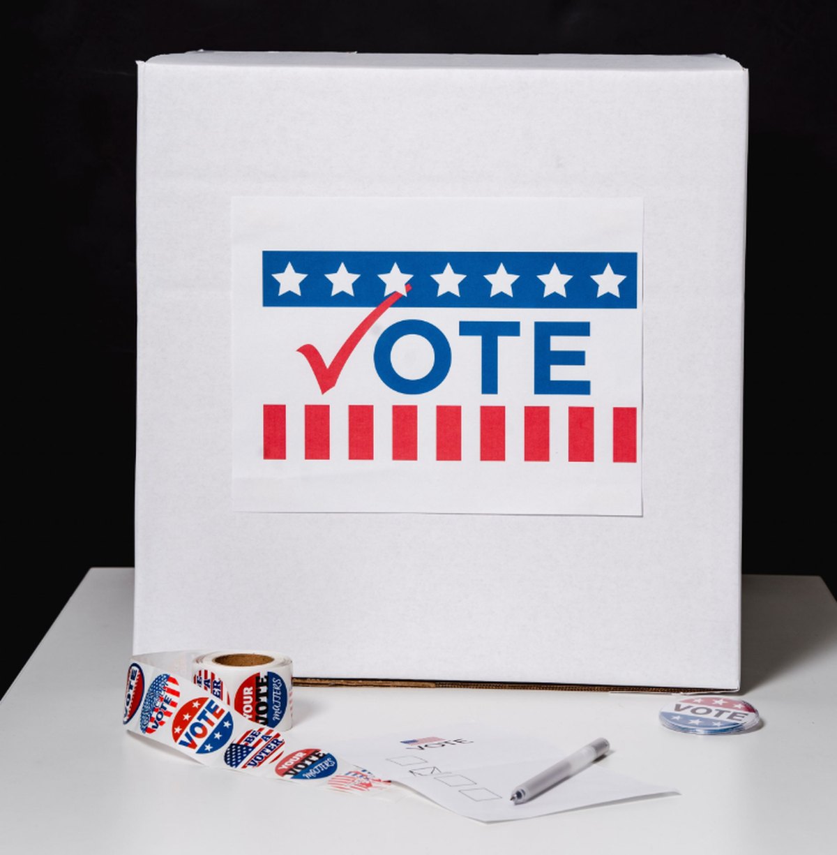 A ballot box marked VOTE with voter stickers, a ballot and a pen on a table.